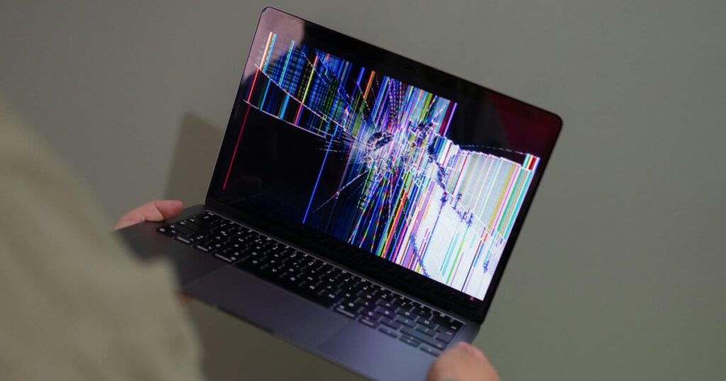 A broken laptop screen displayed with colorful glitch being held by a person.