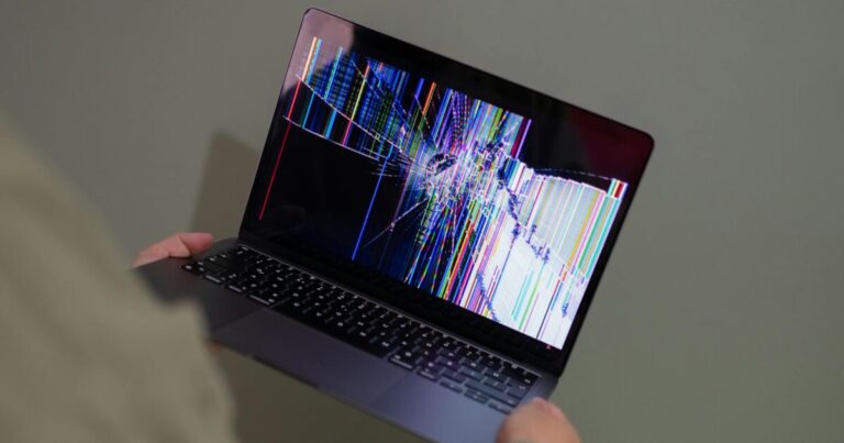 A broken laptop screen displayed with colorful glitch being held by a person.
