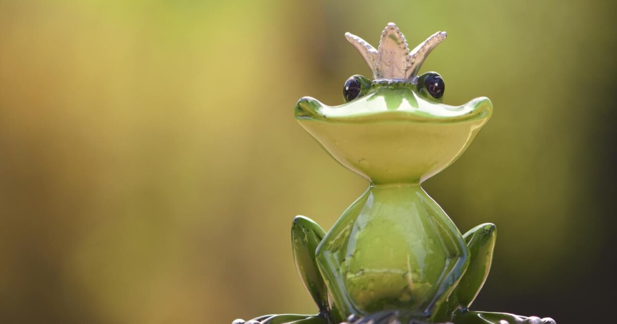 A cute frog prince figurine sits gracefully outdoors with a blurred green background.