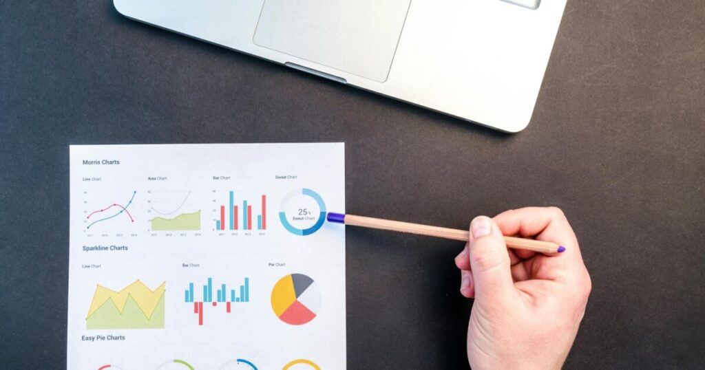 Hand holding pencil reviewing colorful data charts on desk with laptop.