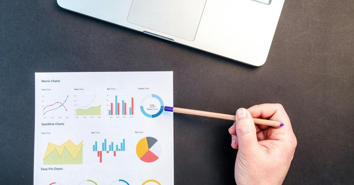 Hand holding pencil reviewing colorful data charts on desk with laptop.